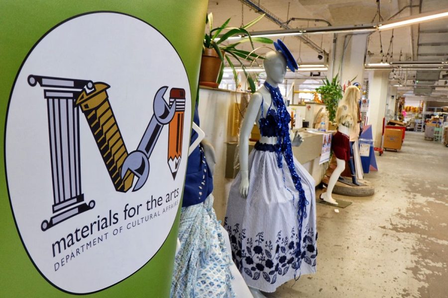 Close up of the green MFTA banner in the MFTA warehouse. The banner displays the MFTA logo with the words "Materials for the Arts Department of Cultural Affairs" beneath. Dressed up mannequins are to the right of the sign.