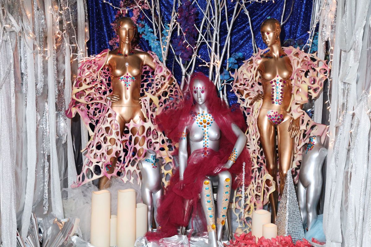 Two bronze and one silver mannequin stand bedazzled with jewels and fabric. Behind them is blue draped fabric and cut strips of silver flow on either side of them. Fairy lights and tree branches are in the background. Large candles rest at the mannequins' feet.