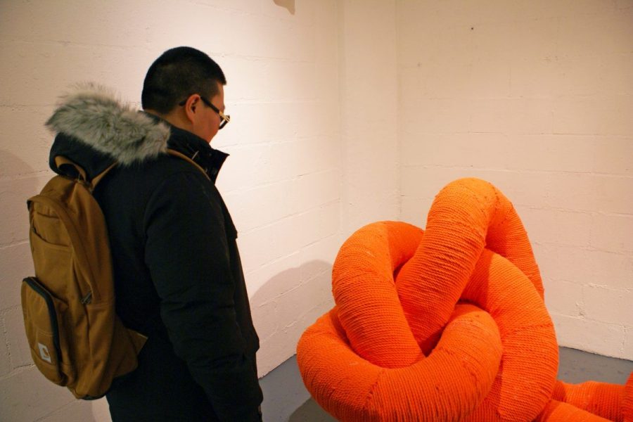 Woman looks at Whitney Oldenburg's sculpture titled Shoe Lace in the MFTA Gallery. The sculpture is made of intertwined ventilation tubing covered in bright orange yarn-like cord.
