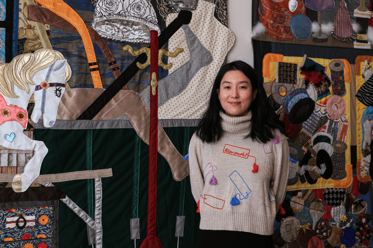 A photo of Woomin Kim standing in front of her large-scale textile works featuring images of MFTA supplies.
