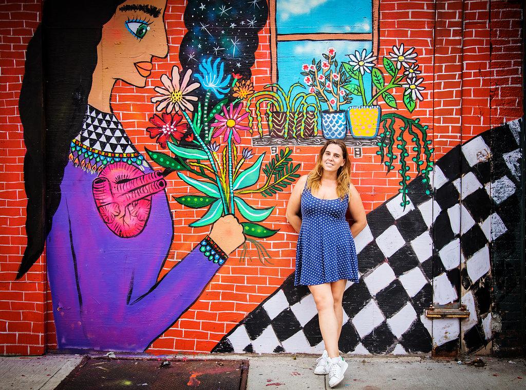 Magda Love stands in front of a mural of a woman holding a flower on a brick building.
