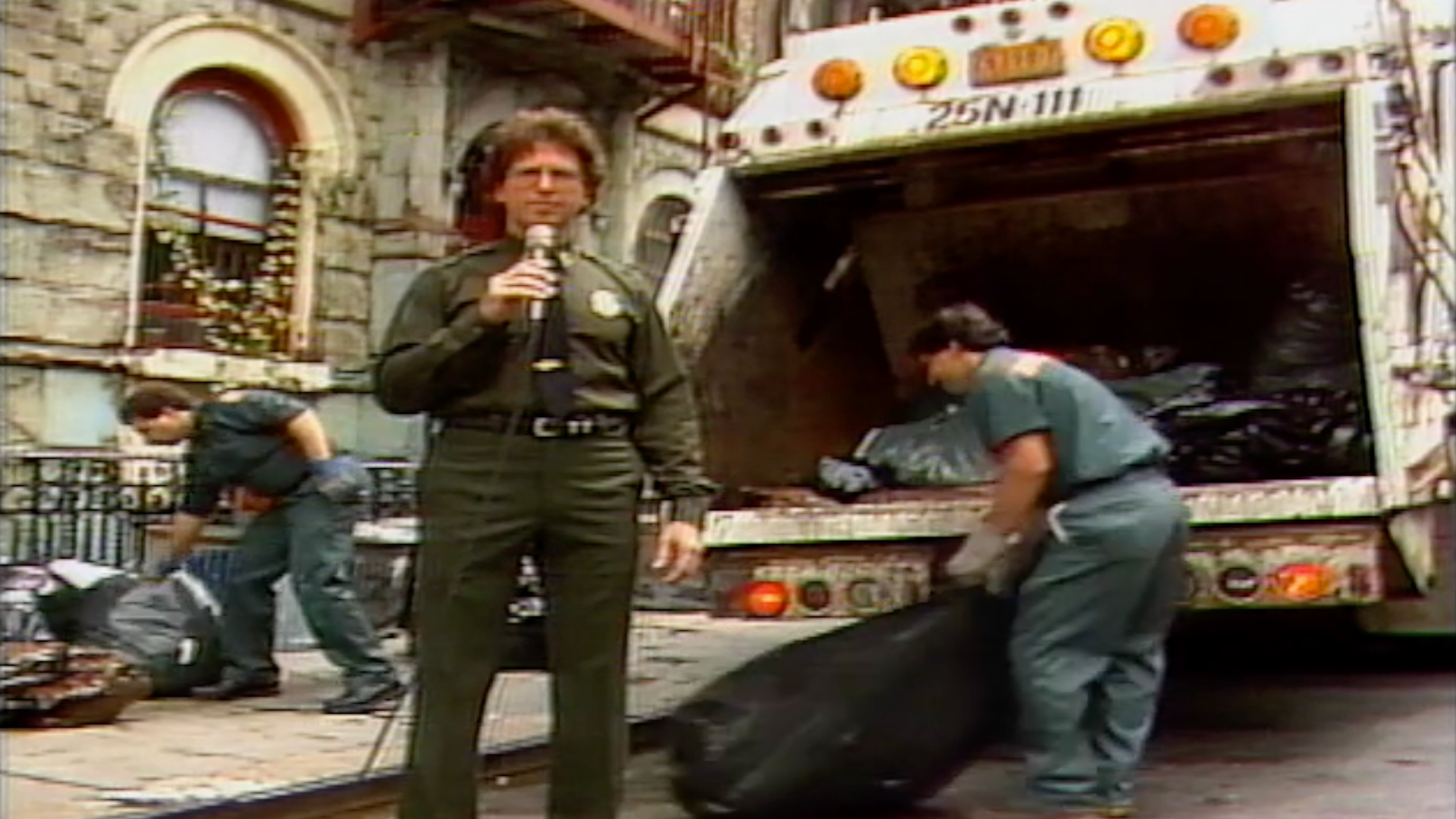 A screenshot of a man with a microphone in front of a Sanitation truck.